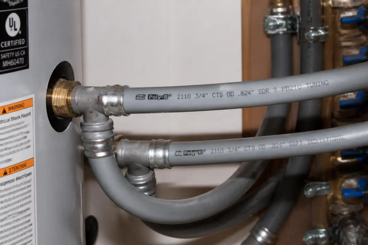Close-up view of gray Poly-B plumbing pipes connected to a water heater, with labels and warning stickers visible on the heater.