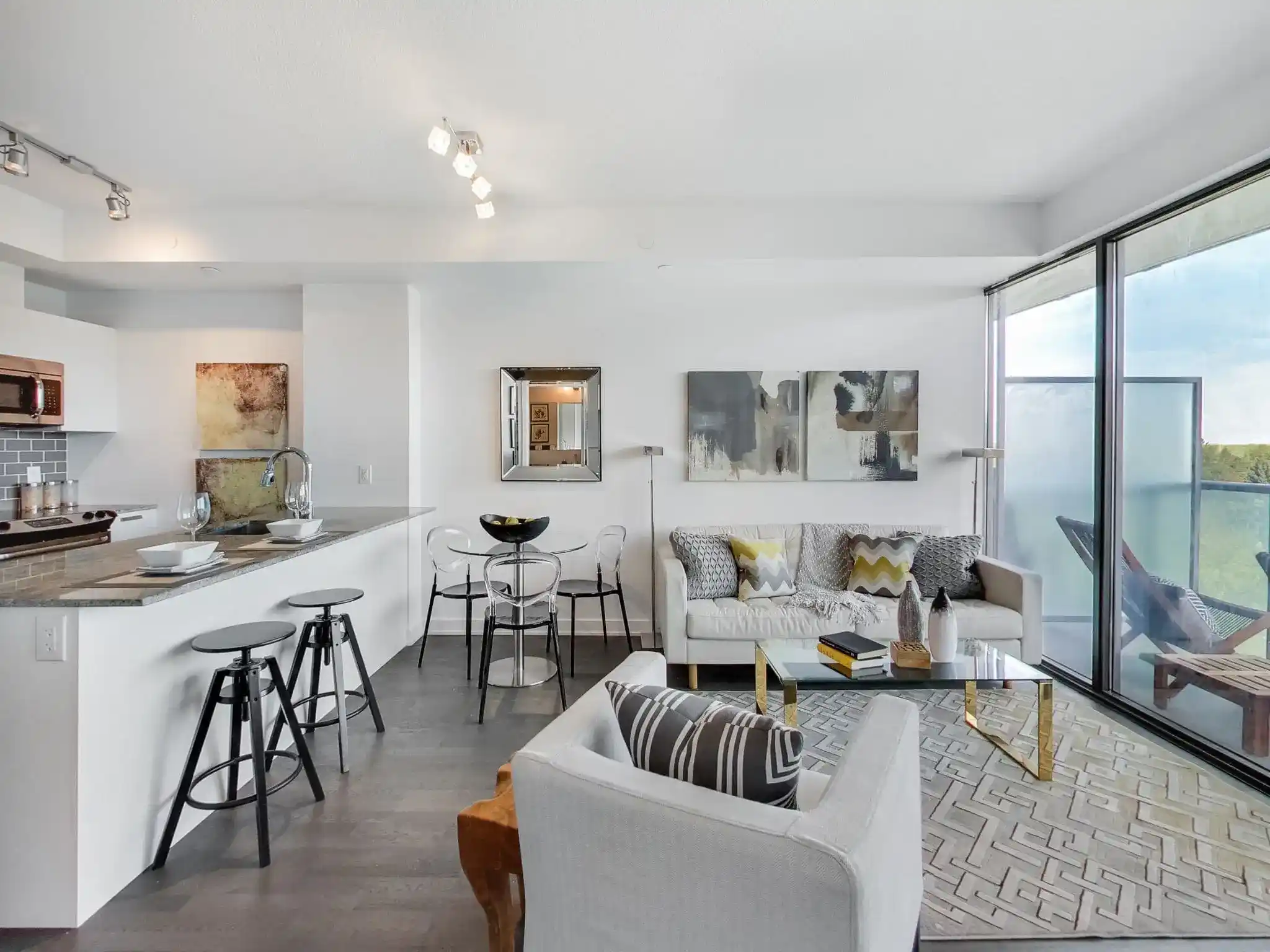 Modern open-plan living room and kitchen with white walls, contemporary furniture, glass coffee table, bar stools, and large windows leading to a balcony with outdoor seating and a view—perfectly showcasing stylish condo & unit interior renovations.