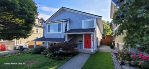 A two-story gray house with a red front door, white trim, and front steps showcases excellent property maintenance, featuring a well-kept lawn and is situated between two neighboring homes.
