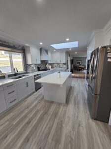 Modern kitchen with white cabinets, a large island, stainless steel appliances, gray backsplash, and wood-look flooring under a skylight—meticulously cared for with regular property maintenance.