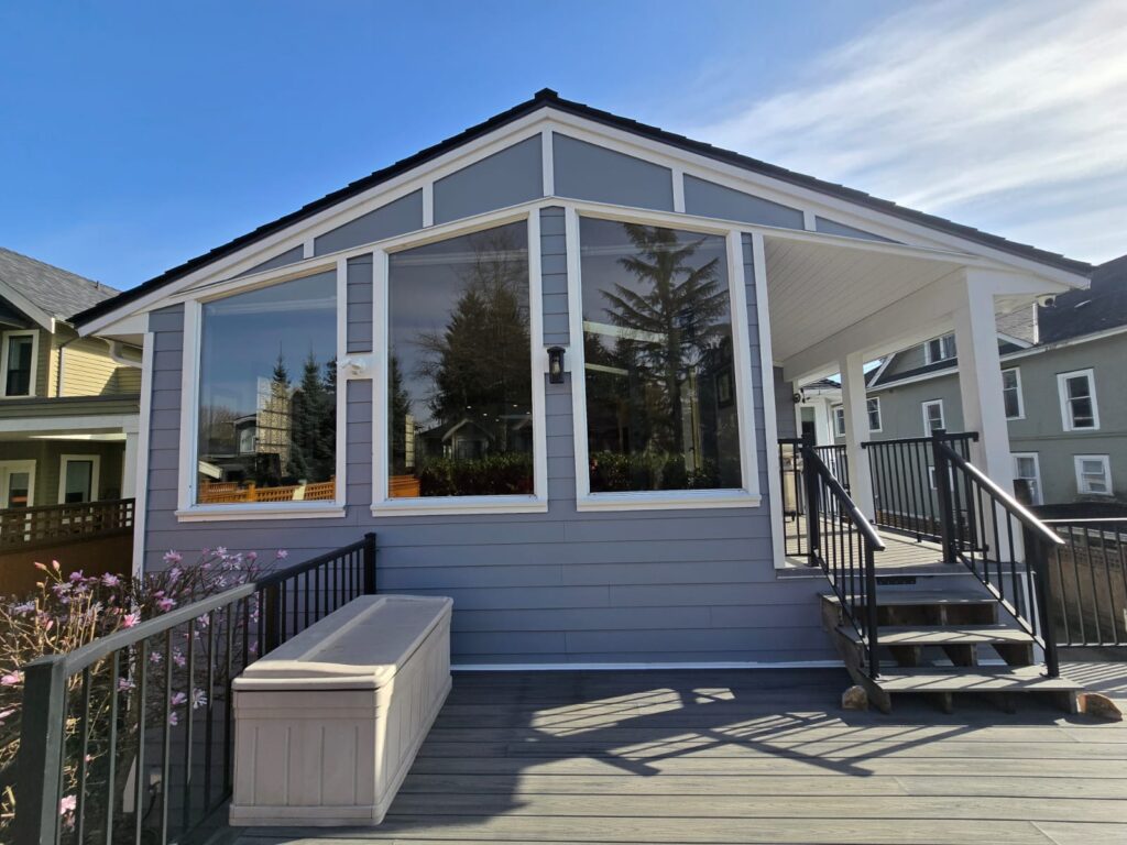 A modern enclosed porch with large windows, gray siding, a white roof, black railings, and a storage bench on a wooden deck—designed with property maintenance in mind—nestled among neighboring houses.