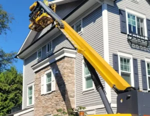 A yellow telescopic boom lift extends up to the roof of a gray house with white trim and stonework on the lower level.