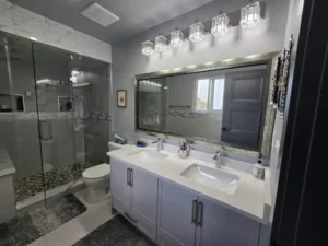 Modern bathroom with double sink vanity, large mirror, overhead lights, glass-enclosed shower, and gray accents.