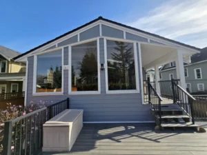 A modern porch with large windows, gray siding, black railings, steps, and a storage bench, attached to a house with trees and neighboring homes visible in the background.