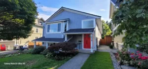 A two-story house with blue-gray siding, white trim, and a red front door. The yard is landscaped with bushes and a concrete walkway leads to the entrance.