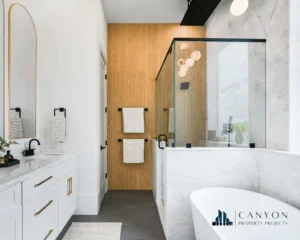 Modern bathroom by expert Bathroom Renovation Contractors featuring a glass-enclosed shower, freestanding bathtub, white vanity with gold accents, wood accent wall, and towel bars; Canyon Property Projects logo in corner.
