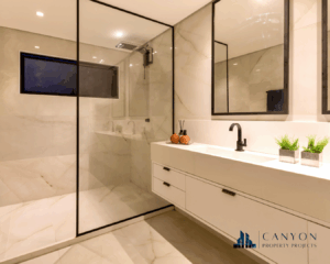 Modern bathroom with a glass-enclosed shower, white marble walls and floor, a long white vanity with black fixtures, and decorative plants; Canyon Property Projects logo in the corner.