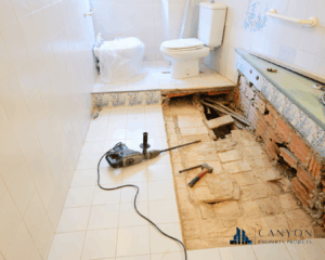 Bathroom under renovation with part of the floor removed, exposing pipes and bricks; tools and construction materials are visible on the tiled floor.