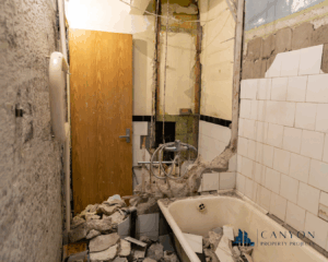 A bathroom mid-renovation with exposed pipes, broken tiles, rubble on the floor, and missing wall sections. A wooden door and bathtub are visible.