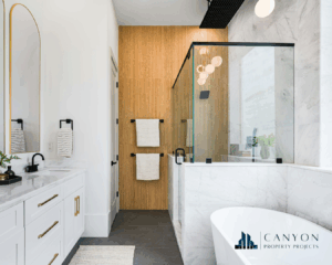Modern bathroom with a glass shower, marble tub, double vanity, gold accents, and wood accent wall with two towel racks. Canyon Property Projects logo in the corner.