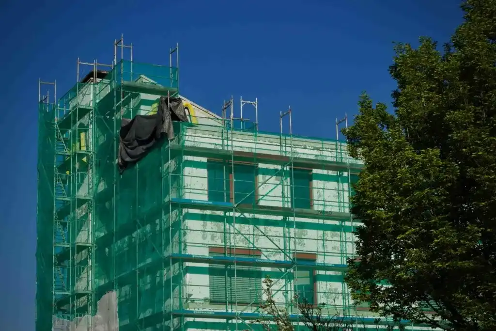 A commercial building under construction is covered in green netting and scaffolding, with a tree in the foreground and a clear blue sky in the background.