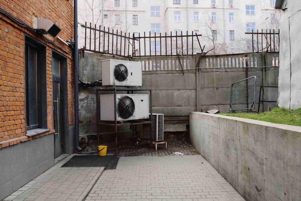 Two outdoor air conditioning units, part of apartment building HVAC upgrades for energy savings, are mounted on a metal frame next to a brick building, with a concrete wall and bent metal fence behind them.