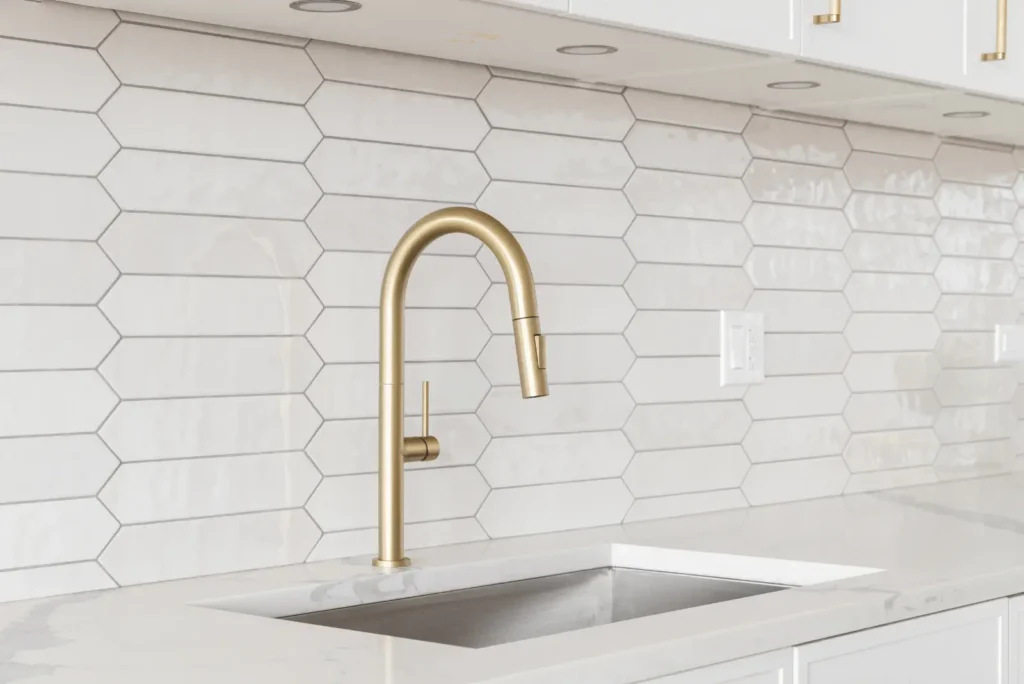 A modern kitchen sink with a gold faucet set against a white hexagonal tile backsplash and white countertop.