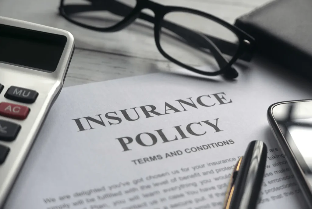 A close-up of an insurance policy document on a desk, surrounded by a pair of eyeglasses, a calculator, a pen, and a smartphone.