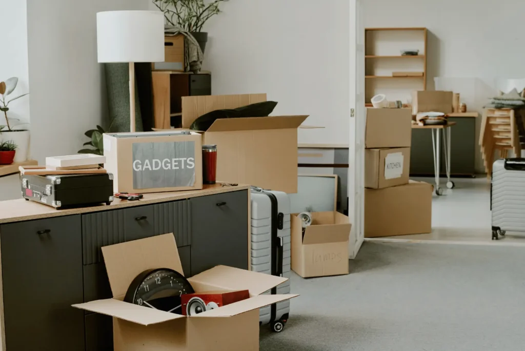 A room filled with cardboard boxes, suitcases, and household items packed for moving, with labels such as "GADGETS," "KITCHEN," and "LAMPS" visible on some boxes.