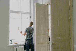 A person in work clothes installs or repairs a window in a room with covered doors and construction tools nearby, showcasing a Step-by-Step Guide to Renovating Older Multifamily Properties.