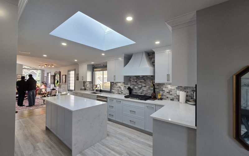 Modern kitchen with white cabinets, marble island, and a skylight; two people are seen standing in the adjacent living area, highlighting the benefits of attentive property maintenance.